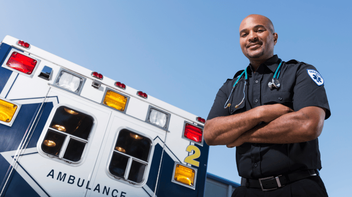 A confident paramedic stands with arms crossed in front of an ambulance, wearing a black uniform with a stethoscope around his neck. The emergency vehicle, marked with flashing lights and labeled "AMBULANCE," is parked under a clear blue sky.