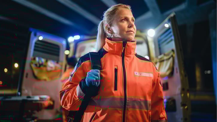A paramedic is standing outside of an ambulance holding a backpack over one shoulder and looking into the distance